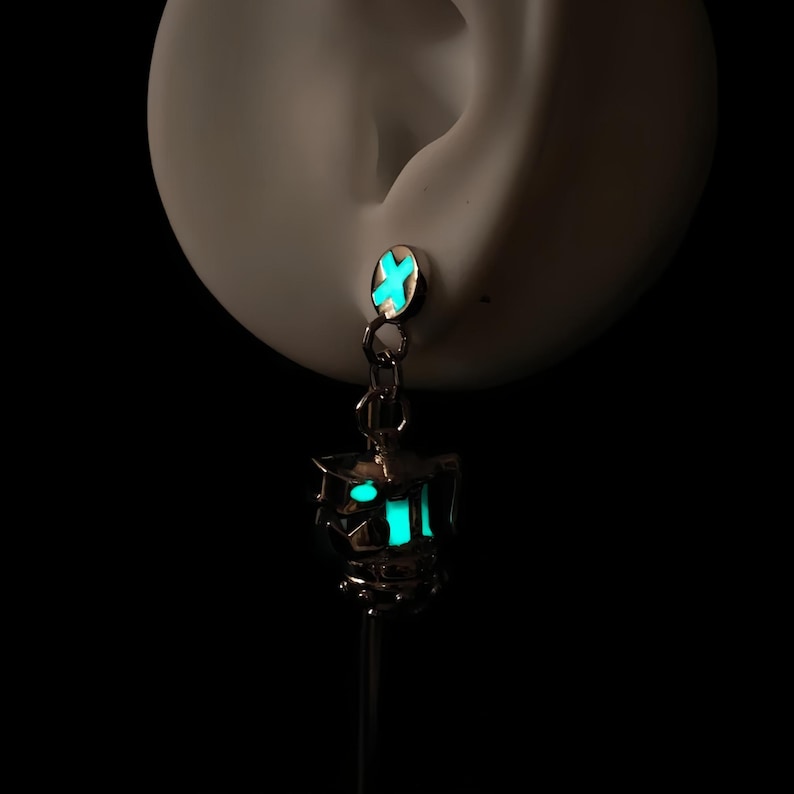 May include: A silver earring with a glow-in-the-dark charm shaped like a lantern. The charm is a teal color and has a silver chain attached to it.