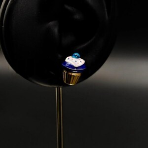 Puede incluir: Un charm de cupcake azul y blanco con base dorada. El cupcake tiene glaseado blanco y chispas azules. El charm est&aacute; sobre un fondo negro.