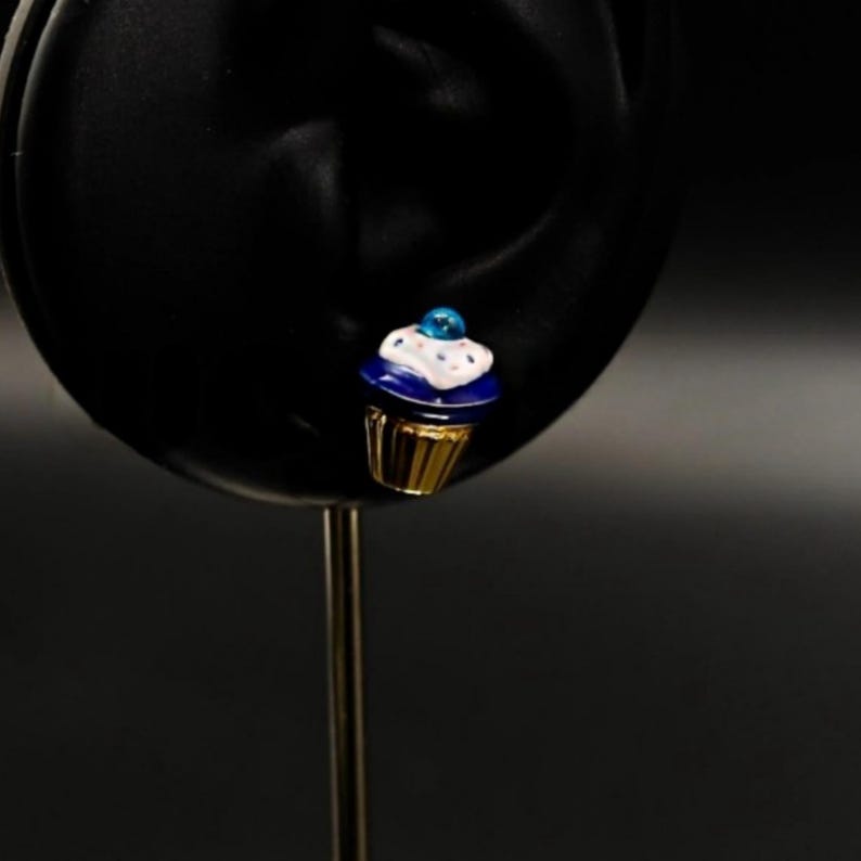 Puede incluir: Un charm de cupcake azul y blanco con base dorada. El cupcake tiene glaseado blanco y chispas azules. El charm est&aacute; sobre un fondo negro.