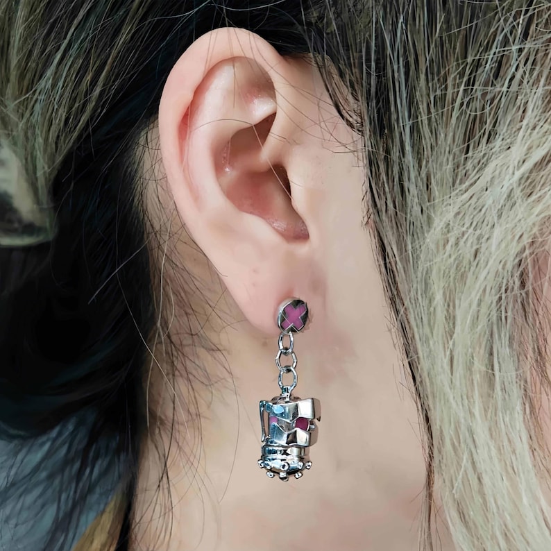 May include: Silver dangle earrings with a pink cross charm and a silver crown charm. The earrings are hanging from a person's ear.