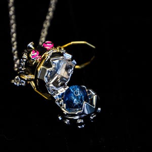 May include: A silver and gold pendant necklace with a detailed, mechanical-style design. The pendant opens to reveal a blue orb. The necklace features pink accents and hangs on a delicate chain against a black background.