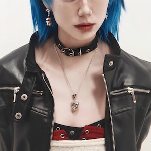 May include: A woman with blue hair wearing a black leather jacket, a black choker with silver buckles, and a silver necklace with a pendant.