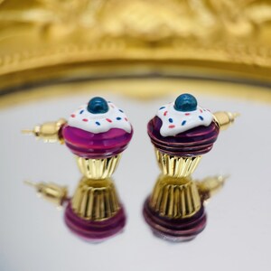Puede incluir: Un par de pendientes de cupcake dorados con glaseado rosa, glaseado blanco, chispas azules y un ar&aacute;ndano azul en la parte superior.