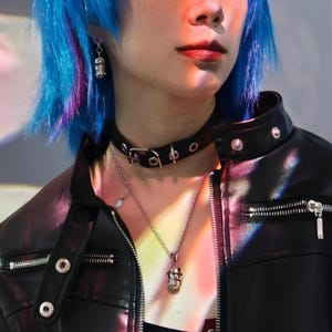 May include: A person with blue hair wearing a black leather jacket with silver hardware. They are wearing a silver chain necklace with a skull pendant and a black choker with silver hardware.