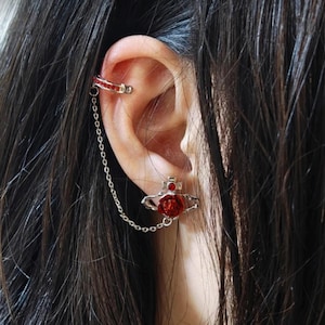 Op de afbeelding: Een zilveren oorcuff met een rode roos bedel en een hangende ketting. De ketting is bevestigd aan een kleine rode roos bedel met een zilveren setting.