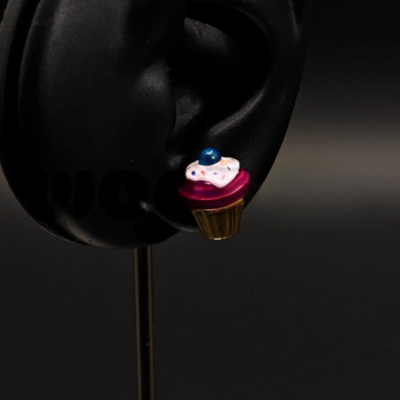 Puede incluir: Un solo pendiente con un peque&ntilde;o cupcake con glaseado rosa, chispas blancas y un ar&aacute;ndano azul en la parte superior. El cupcake es de color dorado en la parte inferior.