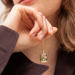 May include: A gold necklace with a rectangular pendant. The pendant is clear with a gold frame and contains green and pink floral elements. The necklace chain is also gold and has small diamond accents.