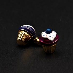Puede incluir: Dos abalorios de cupcake de color dorado con detalles de esmalte de colores. Un cupcake es morado con glaseado blanco y el otro es rojo con glaseado blanco y una chispa azul en la parte superior.