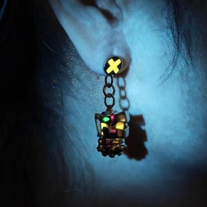 May include: A silver earring with a yellow and green charm shaped like a robot head. The charm is attached to a silver chain with a yellow and black cross charm at the top.