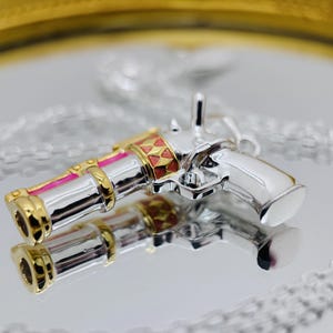 May include: A silver-colored pendant necklace featuring a detailed gun-shaped charm. The charm has gold-colored accents, pink stripes, and red and gold geometric patterns. The pendant is reflected in the shiny surface below.
