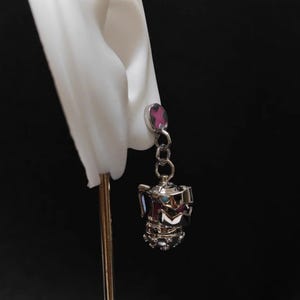 May include: Silver dangle earrings with a pink cross charm and a silver teapot charm.