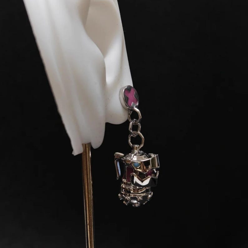 May include: Silver dangle earrings with a pink cross charm and a silver teapot charm.