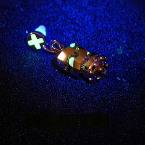May include: A glow-in-the-dark charm featuring a detailed robot design with a crown on top. The charm is attached to a small, round charm with a plus sign.