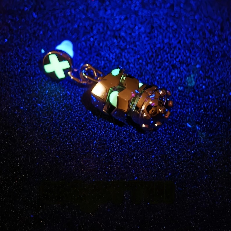 May include: A glow-in-the-dark charm featuring a detailed robot design with a crown on top. The charm is attached to a small, round charm with a plus sign.
