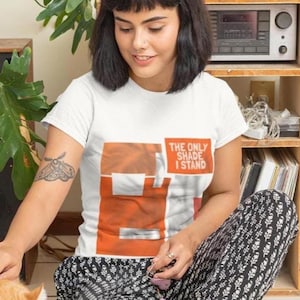 Könnte beinhalten: Weißes T-Shirt mit dem Text "THE ONLY SHADE I STAND" in Orange und Weiß. Das Shirt hat ein orangefarbenes Grafikdesign. Die Person trägt eine Hose mit schwarz-weißem Muster.