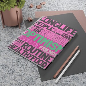 Könnte beinhalten: Ein spiralgebundenes Notizbuch mit rosa Einband, auf dem die Wörter "Optimismus", "Langes Leben" und "Gesunde Ernährung" stehen. Zwei Bleistifte, einer braun und einer weiß, liegen neben dem Notizbuch auf einer grauen Oberfläche.