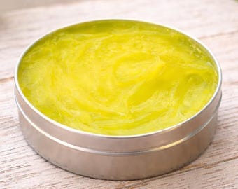 May include: A round, silver-colored metal tin filled with a translucent, bright yellow gel substance. The gel has a smooth, swirled texture that fills the container to the brim, creating a vibrant contrast against the metallic rim of the tin.