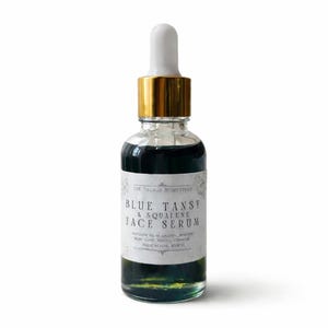 Pu&ograve; includere: Un flacone contagocce in vetro trasparente contenente un liquido blu scuro. L'etichetta bianca riporta la scritta The Village Homestead Blue Tansy & Squalene Face Serum. Il flacone presenta un collare dorato e un tappo contagocce bianco su sfondo bianco.