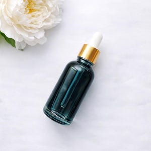 Pu&ograve; includere: Un flacone contagocce in vetro color verde ottanio scuro con un collare dorato e un bulbo in gomma bianca, posizionato accanto a una grande peonia bianca a petali multipli. Il flacone &egrave; cilindrico con una superficie liscia e riflettente.