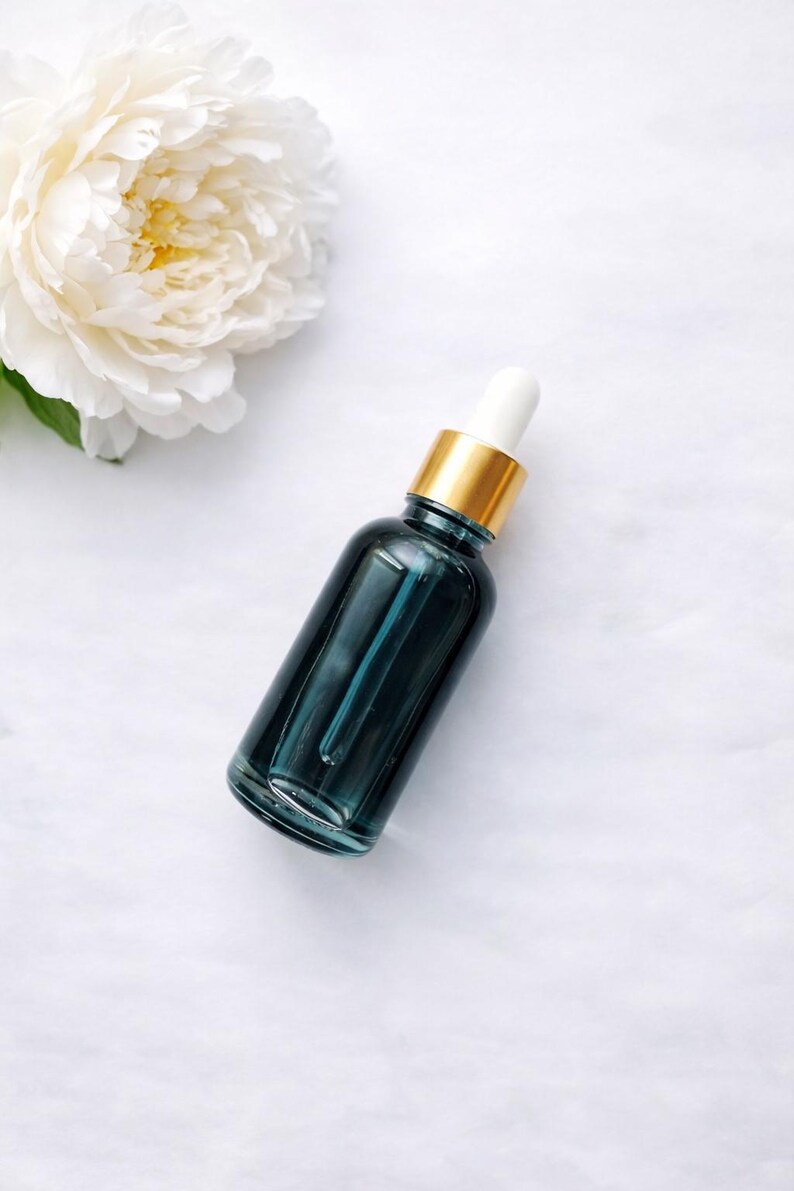 Pu&ograve; includere: Un flacone contagocce in vetro color verde ottanio scuro con un collare dorato e un bulbo in gomma bianca, posizionato accanto a una grande peonia bianca a petali multipli. Il flacone &egrave; cilindrico con una superficie liscia e riflettente.