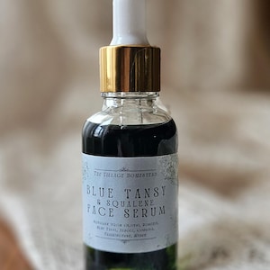 Pu&ograve; includere: Un flacone contagocce in vetro trasparente contenente un liquido verde scuro. L'etichetta riporta The Village Homestead Blue Tansy & Squalene Face Serum, con testo aggiuntivo che elenca ingredienti come olio d'oliva, rosmarino, tanaceto, olivello spinoso e copaiba.