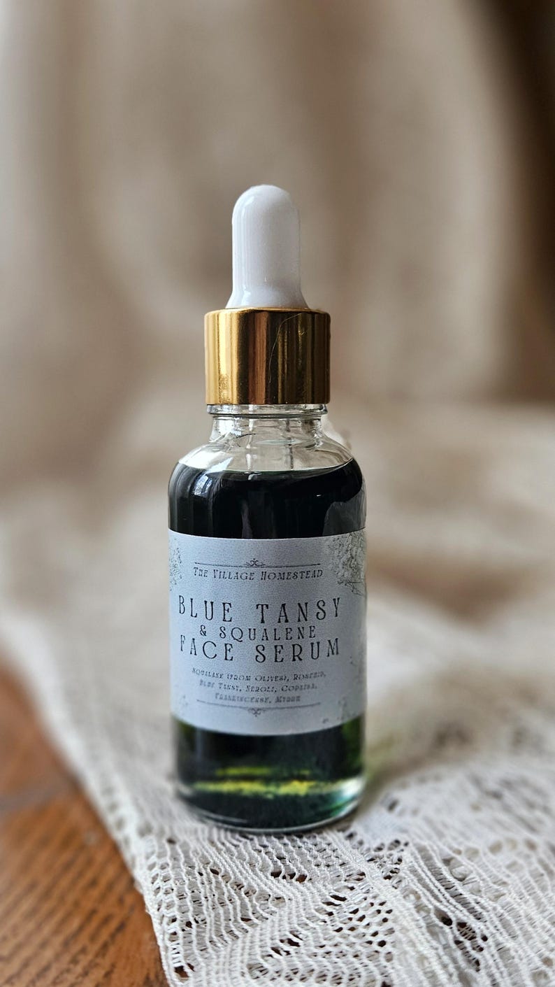 Pu&ograve; includere: Un flacone contagocce in vetro trasparente contenente un liquido verde scuro. L'etichetta riporta The Village Homestead Blue Tansy & Squalene Face Serum, con testo aggiuntivo che elenca ingredienti come olio d'oliva, rosmarino, tanaceto, olivello spinoso e copaiba.