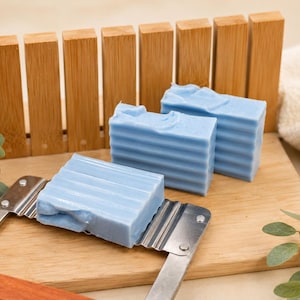 May include: Three rectangular bars of light blue soap with a ribbed texture on the sides. One bar is positioned on a metal soap cutter with a wooden handle, while two other bars are stacked behind it on a wooden surface with bamboo slats.
