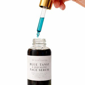 Pu&ograve; includere: Un flacone in vetro trasparente di siero viso Blue Tansy & Squalene con un'etichetta bianca e testo nero. Un contagocce in vetro con colletto dorato e bulbo bianco &egrave; tenuto sopra il flacone aperto, rilasciando una goccia di siero liquido blu.
