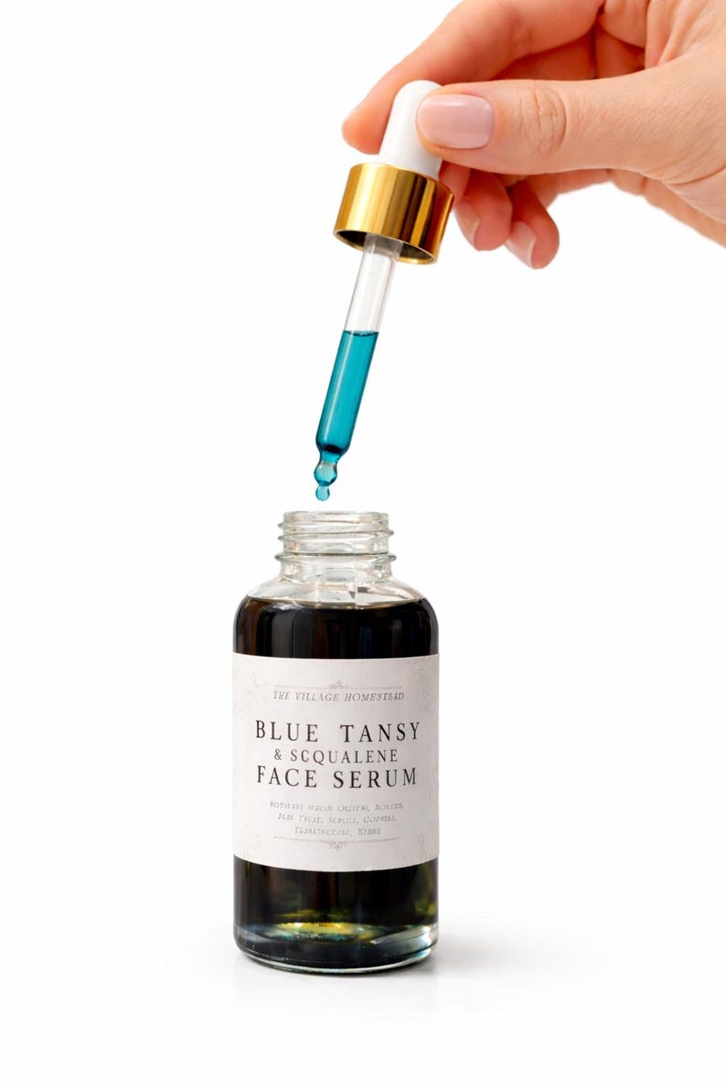 Pu&ograve; includere: Un flacone in vetro trasparente di siero viso Blue Tansy & Squalene con un'etichetta bianca e testo nero. Un contagocce in vetro con colletto dorato e bulbo bianco &egrave; tenuto sopra il flacone aperto, rilasciando una goccia di siero liquido blu.