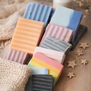 May include: A collection of colorful, rectangular soaps in various shades of blue, pink, orange, and black. Some soaps have a ridged texture. The soaps are arranged on a wooden surface with decorative stars and a knitted fabric.