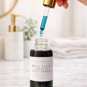 Pu&ograve; includere: Un flacone in vetro trasparente di siero viso Blue Tansy & Squalene con etichetta bianca e testo nero. Un contagocce in vetro con tappo dorato &egrave; tenuto sopra il flacone aperto, rilasciando una goccia di liquido blu vibrante nel siero scuro.