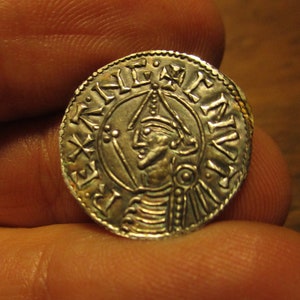 May include: A silver coin with a portrait of a person wearing a crown and holding a scepter. The coin has the inscription "REX" and "ANGLIE" around the edge.