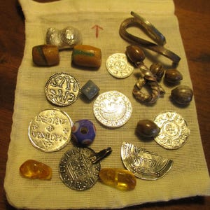 Puede incluir: Una bolsa de tela blanca llena de varios artículos, incluyendo monedas, cuentas y un anillo de metal. Las monedas son de plata y tienen diferentes diseños. Las cuentas son marrones, verdes y azules. El anillo de metal es de plata y tiene un diseño simple.