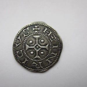 May include: A silver coin with intricate designs. The coin features a central cross design surrounded by circular patterns and text. The edges are detailed with small, repeating shapes. The coin has a worn, antique appearance.