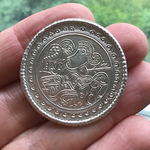May include: A silver coin with an embossed design featuring a figure with ornate headwear and a bird. The coin has a detailed border with small circular patterns. The coin is held in a hand against a blurred green background.