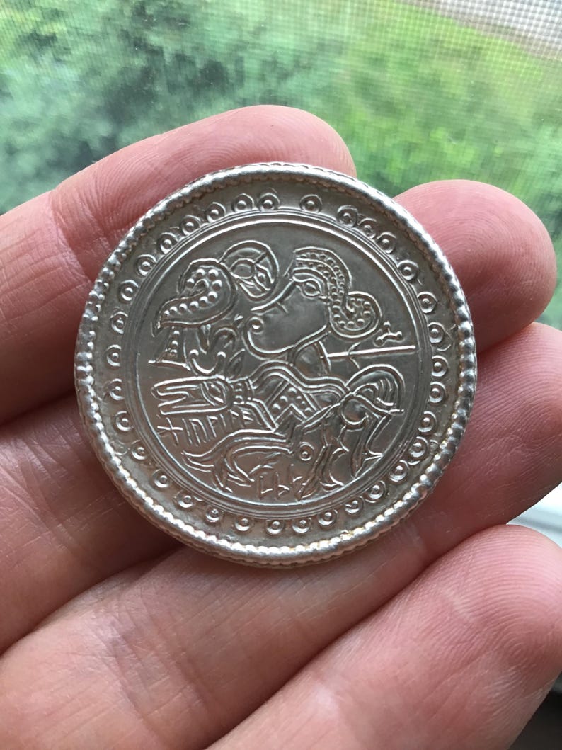 May include: A silver coin with an embossed design featuring a figure with ornate headwear and a bird. The coin has a detailed border with small circular patterns. The coin is held in a hand against a blurred green background.