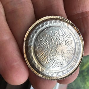 May include: A silver-colored, round metal object with an intricate embossed design. The object has a detailed pattern in the center and a textured border. The item is held in a hand, suggesting its size and portability. The design appears to be of historical or cultural significance.