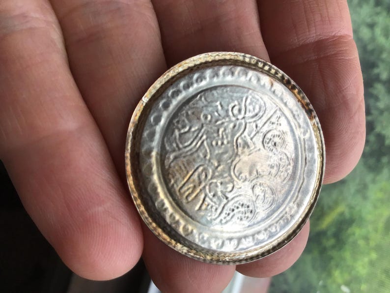 May include: A silver-colored, round metal object with an intricate embossed design. The object has a detailed pattern in the center and a textured border. The item is held in a hand, suggesting its size and portability. The design appears to be of historical or cultural significance.
