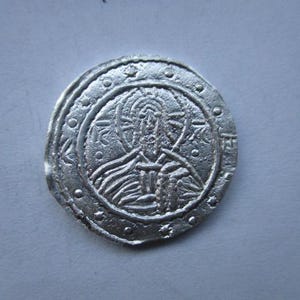 May include: A silver coin with a detailed embossed design. The coin features a central figure surrounded by a circular border with small dots and floral patterns. The design is intricate and appears to be of historical or religious significance.