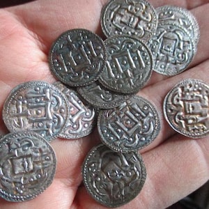 May include: A collection of silver-colored, round coins with intricate embossed designs. The coins are held in a hand, showcasing their detailed patterns and aged appearance. The designs include geometric shapes and stylized figures.