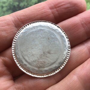 May include: A silver-colored, round metal object with a textured edge. The center of the object is smooth and reflective. The object is held in a hand, with a blurred green background visible.