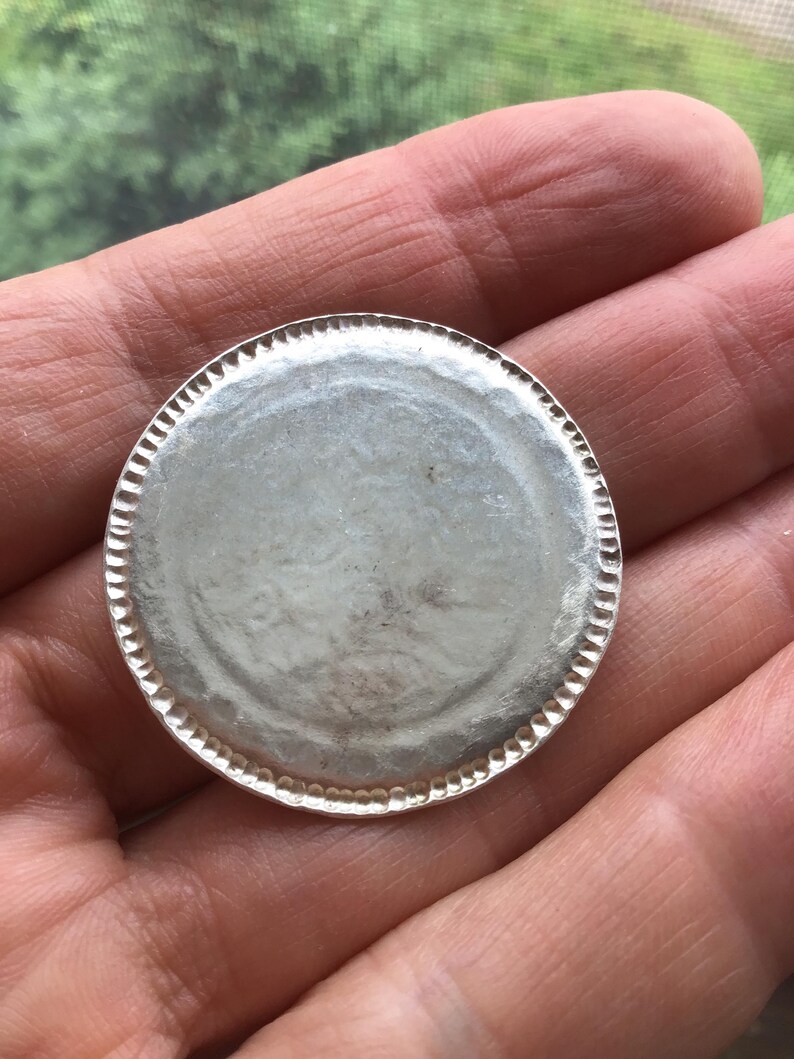 May include: A silver-colored, round metal object with a textured edge. The center of the object is smooth and reflective. The object is held in a hand, with a blurred green background visible.