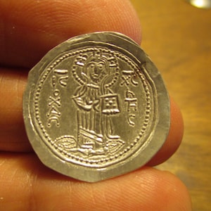 May include: A silver coin with a raised image of a person holding a book. The coin has a beaded border and the inscription "IC XC" around the edge.
