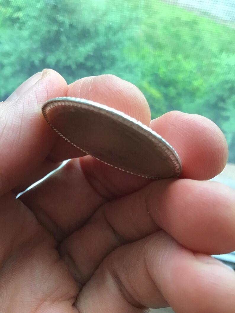 May include: A silver-colored, oval-shaped coin with a detailed, textured edge. The coin is held between fingers, with a blurred green background visible. The coin's surface appears to be a light brown color.