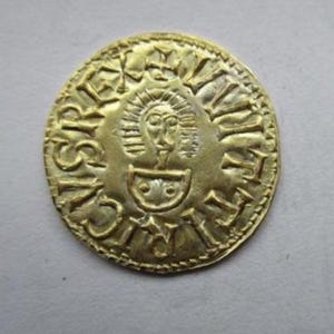 May include: A gold coin with intricate engravings. The coin features a central face design surrounded by text and a cross at the top. The coin's surface has a slightly worn appearance, suggesting age and historical significance.