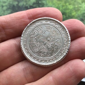 May include: A silver coin with an embossed design featuring a profile and bird-like figures. The coin has a beaded border and is held in a hand. The coin's diameter is approximately 1.5 inches.