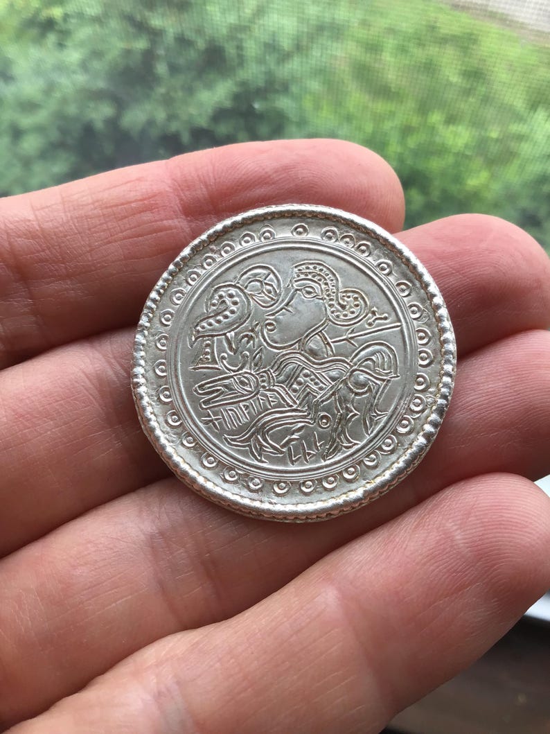 May include: A silver coin with an embossed design featuring a profile and bird-like figures. The coin has a beaded border and is held in a hand. The coin's diameter is approximately 1.5 inches.