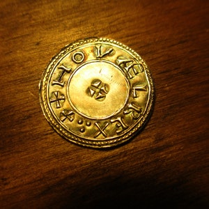 May include: A gold coin with intricate designs and lettering. The coin features a central circle with a cross-shaped design and a ring of lettering around the edge.