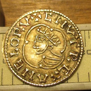 May include: A gold-colored coin with intricate details. The coin features a profile of a person with a crown and inscriptions around the edge. The coin is placed on a ruler, showing a measurement of approximately 20 units.