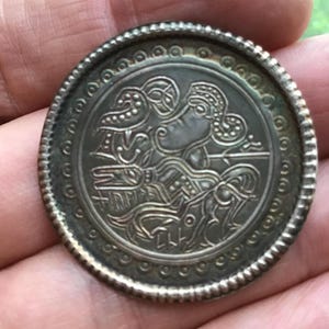 May include: A silver-colored, round coin with an intricate embossed design. The central image features a stylized figure with detailed patterns, surrounded by a decorative border. The coin's edge has a textured, beaded pattern.